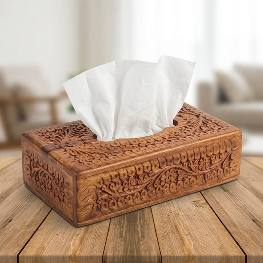 An ornate rectangular wooden tissue box cover with intricate floral carvings, holding white tissue papers, set on a rustic wooden table with a softly blurred living room background.
